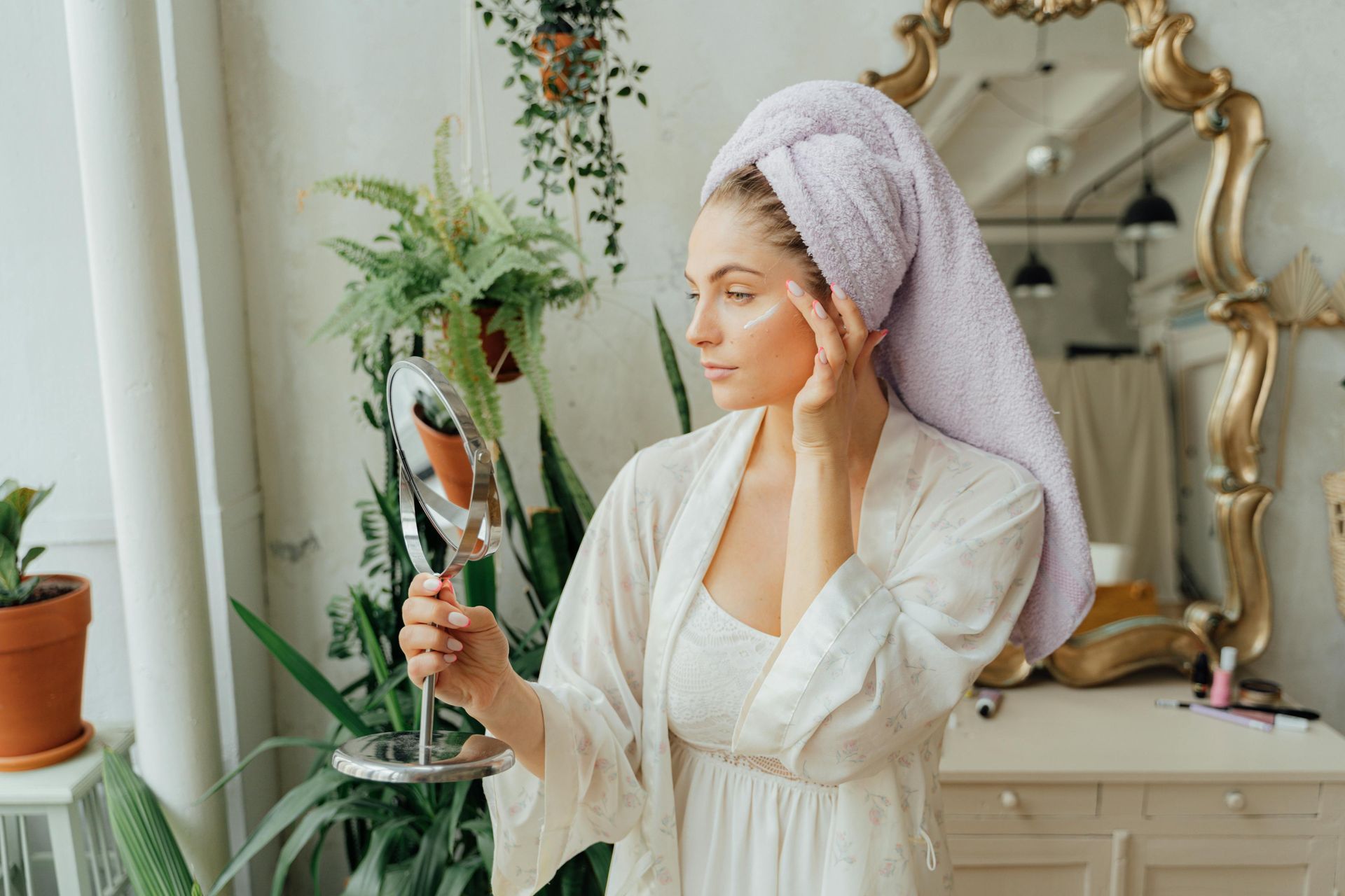 A woman with a towel wrapped around her head is looking at her face in a mirror.