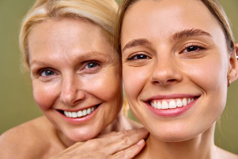 Mother and daughter smiling, close-up. Older woman's arm on younger woman's shoulder; both have fair skin and blonde hair.