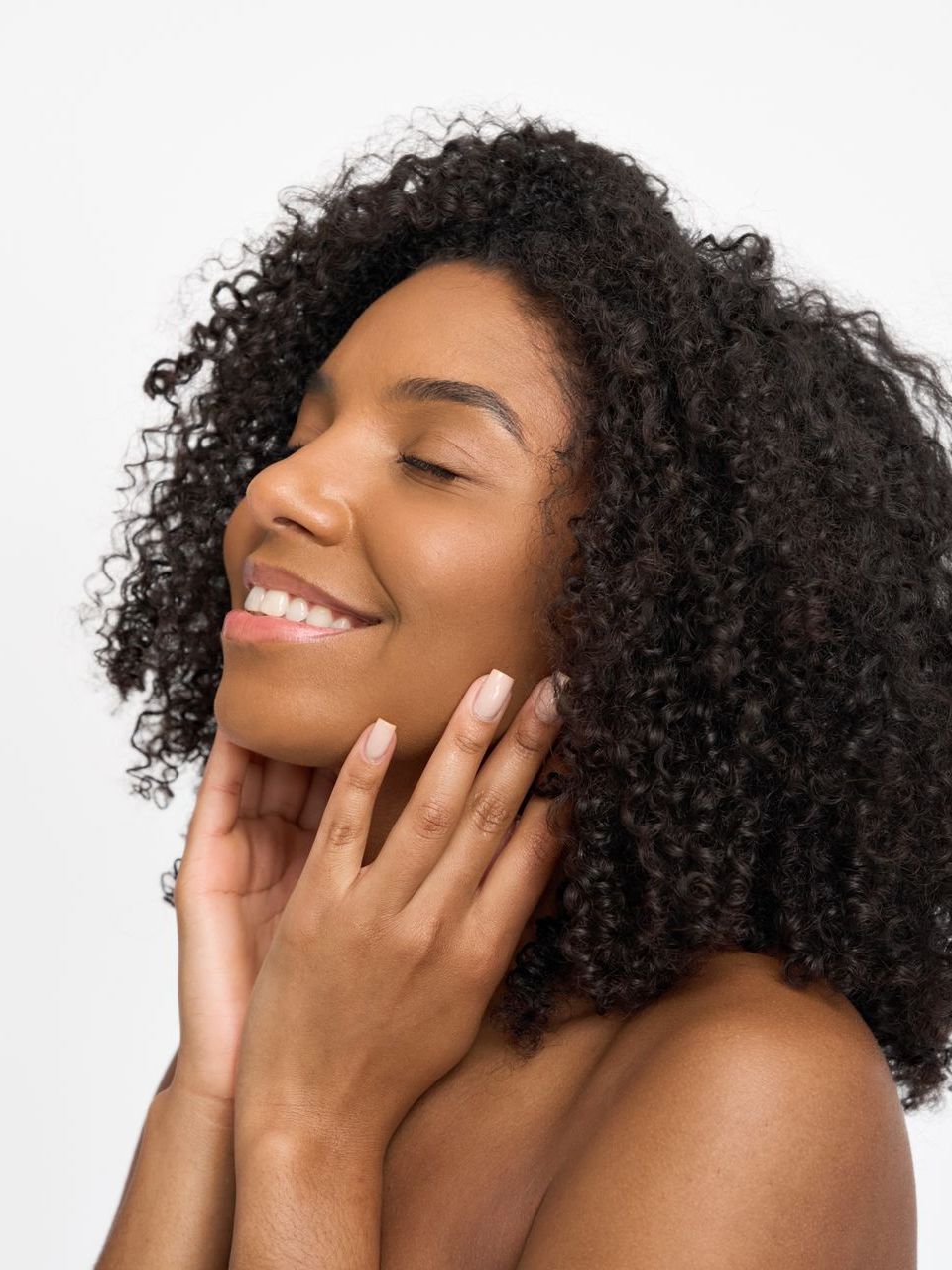 A woman with curly hair is smiling and touching her face.