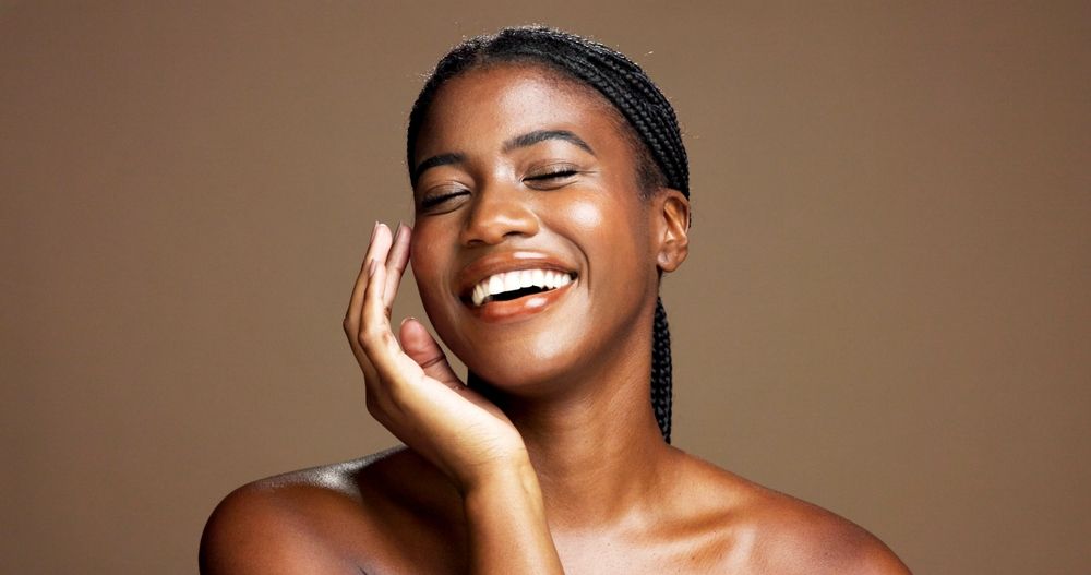 Smiling Black woman with braided hair, eyes closed, touching her face, against a brown background.