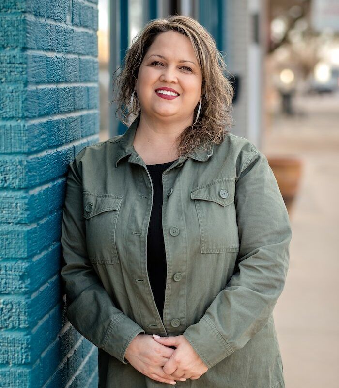 Smiling woman in a green jacket leaning against a blue brick wall outdoors