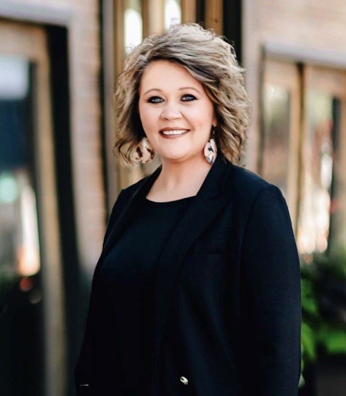 Woman in a black blazer smiling outdoors against a blurred building background