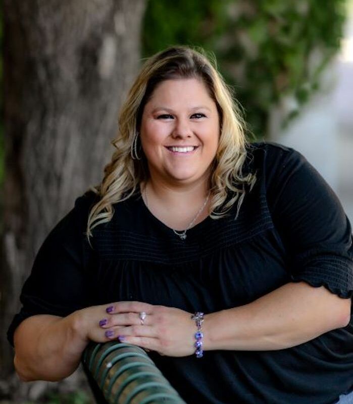Smiling person in a black top leaning on a chair outdoors, with a blurred green background
