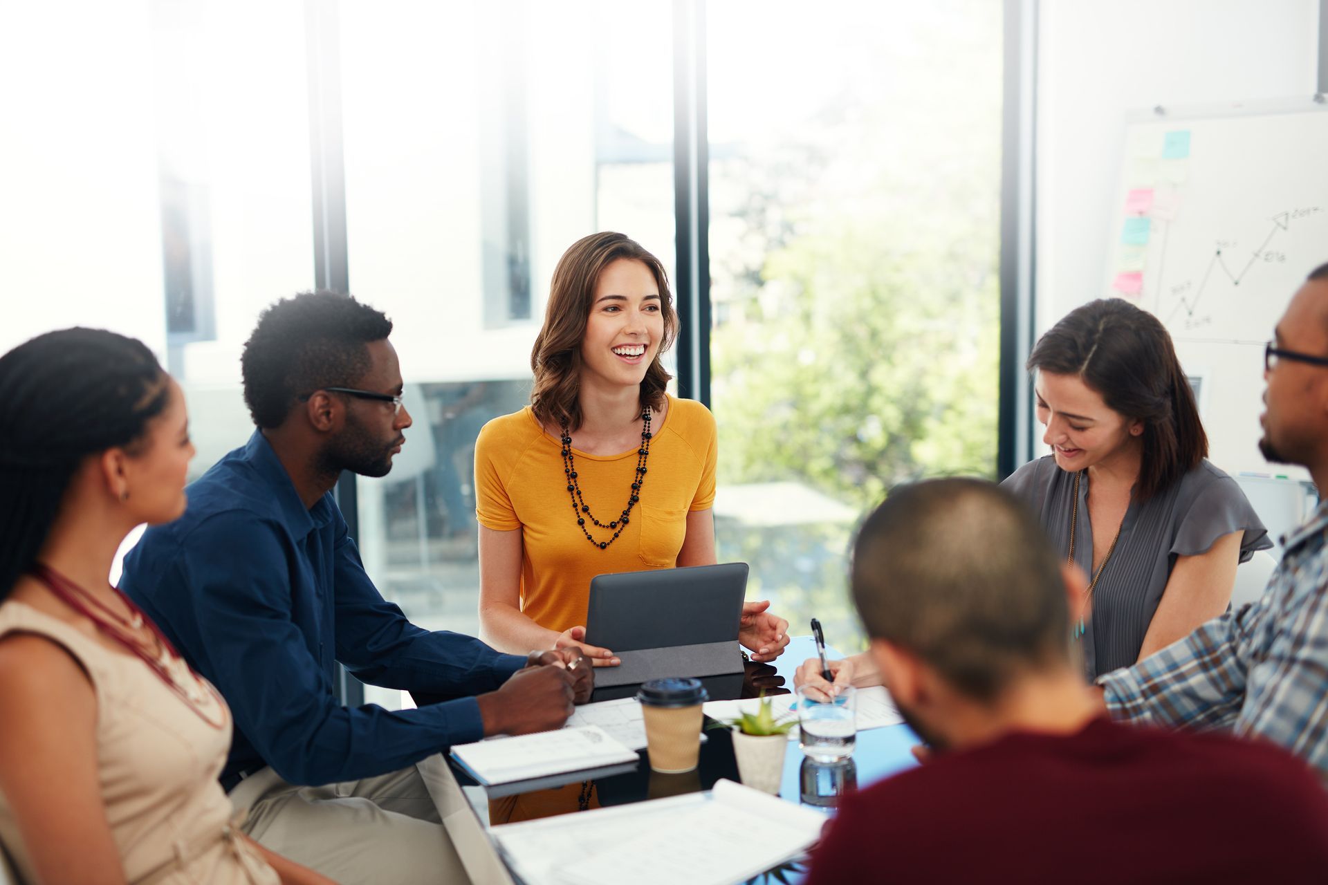 Three businesspeople at a wooden table, reviewing a tablet. Indoor, with plants.