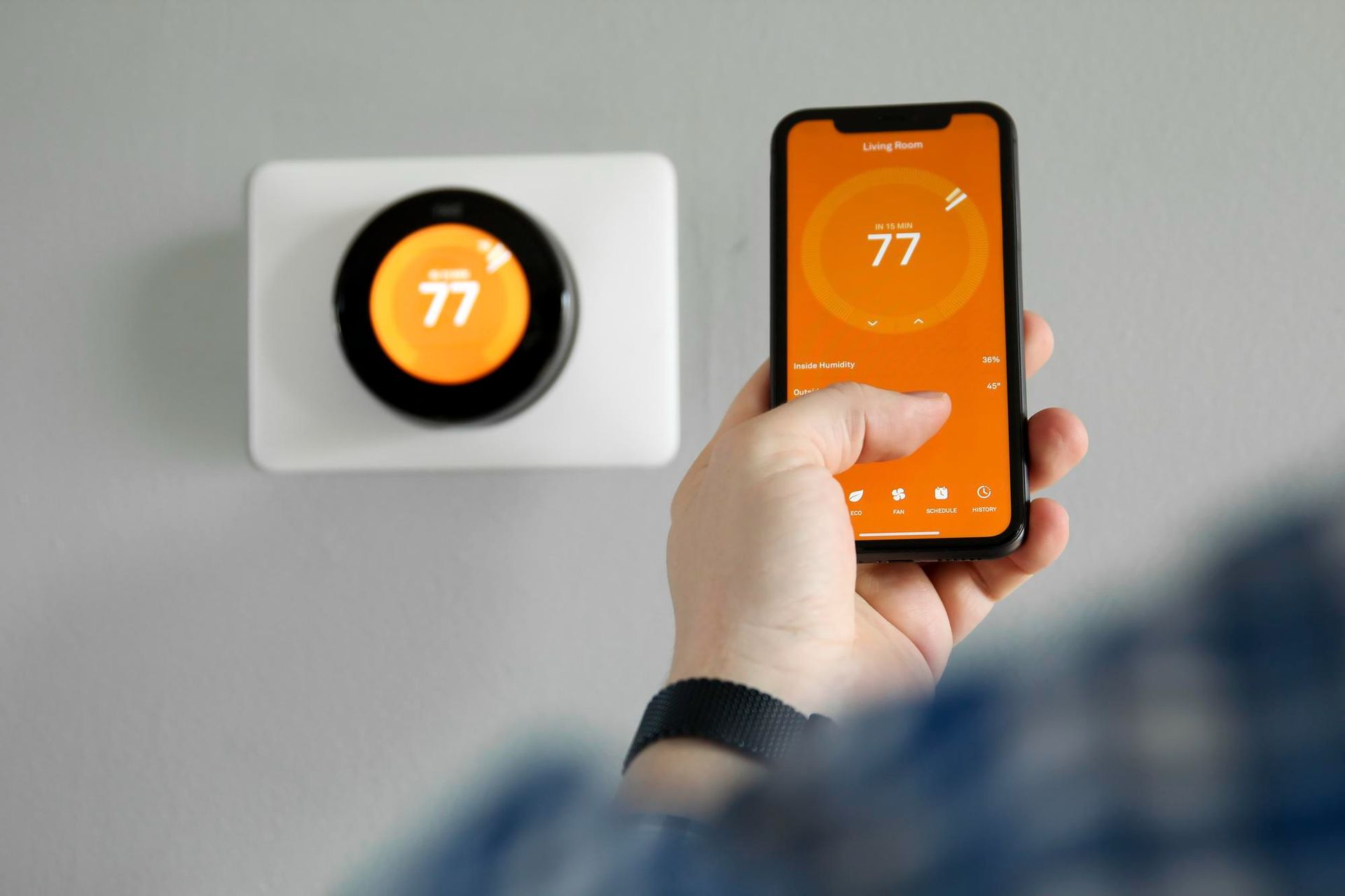 A person uses a smartphone to adjust a Nest smart thermostat set to 77 degrees on a wall.