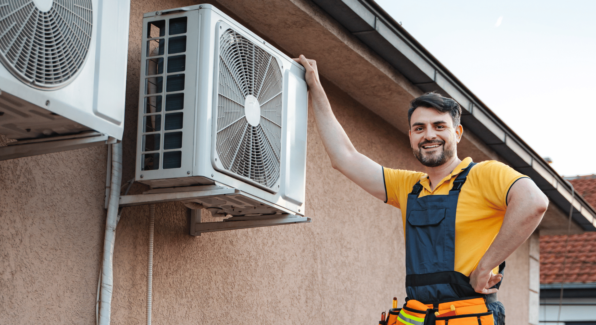 Two technicians in blue uniforms and yellow gloves smile while repairing an outdoor air conditioning unit.