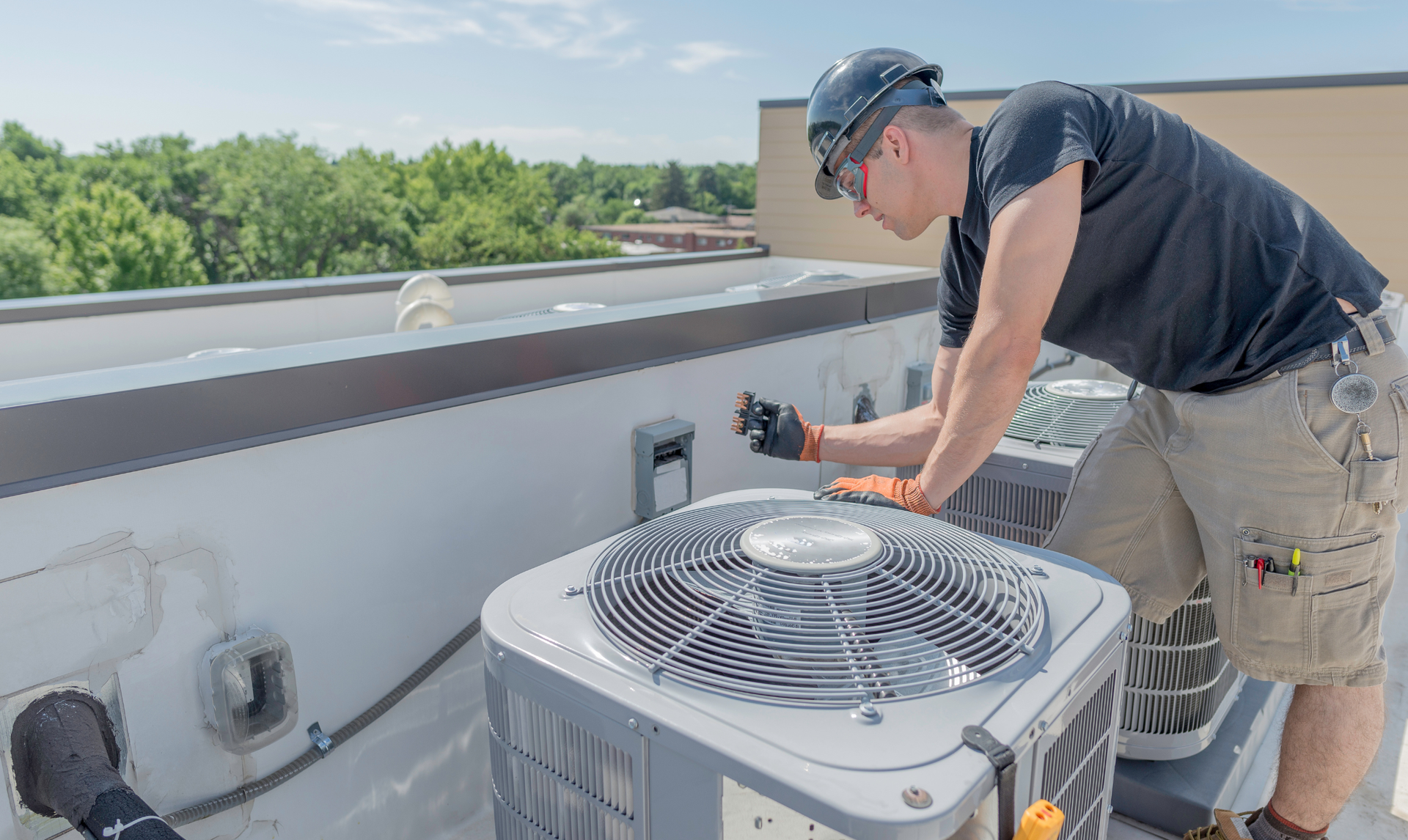 A technician in a hard hat and protective gloves repairs an air conditioning unit on a flat rooftop.