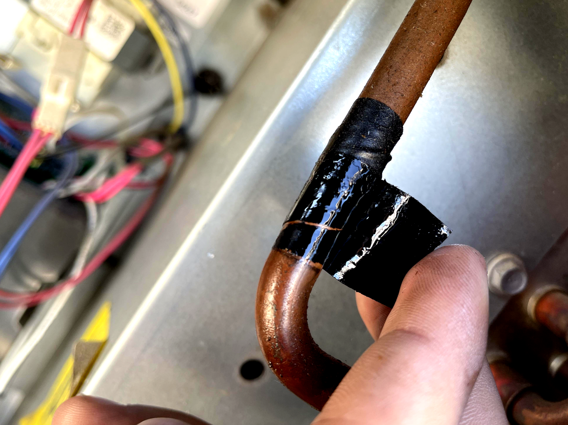 A finger peels black tape off a copper pipe inside an HVAC unit.