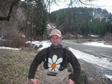 A man wearing a sweater with a flower on it is standing next to a river.