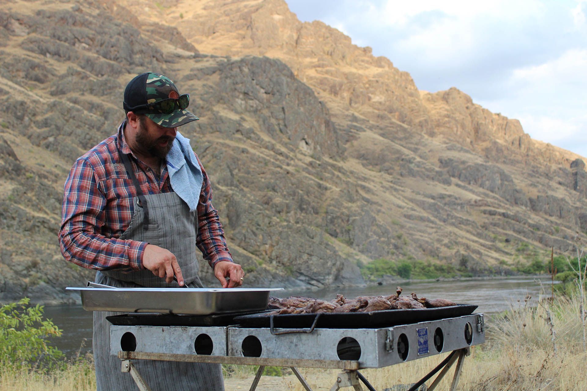 A man is cooking food on a grill in the mountains.