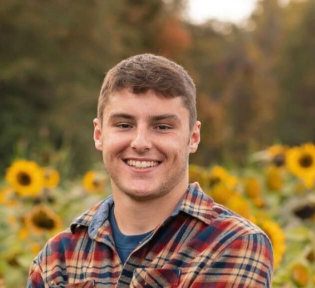 A young man in a plaid shirt is smiling in front of a field of sunflowers.