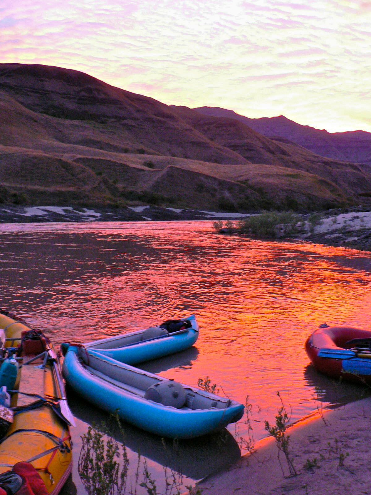 Three kayaks are sitting on the shore of a river at sunset