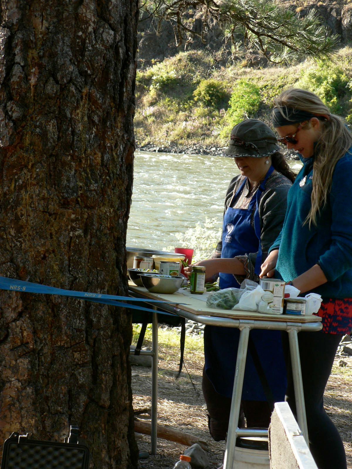 Two women are preparing food in front of a river.