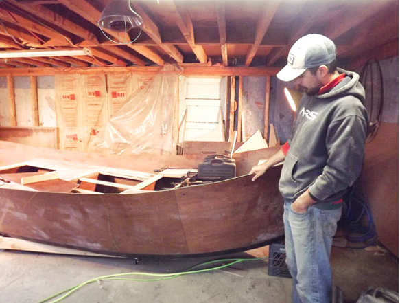 A man wearing a hat and a grey hoodie is standing next to a wooden boat