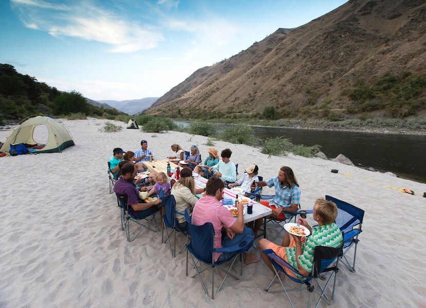 Group of People Sitting Around a Table in The Sand Near a River