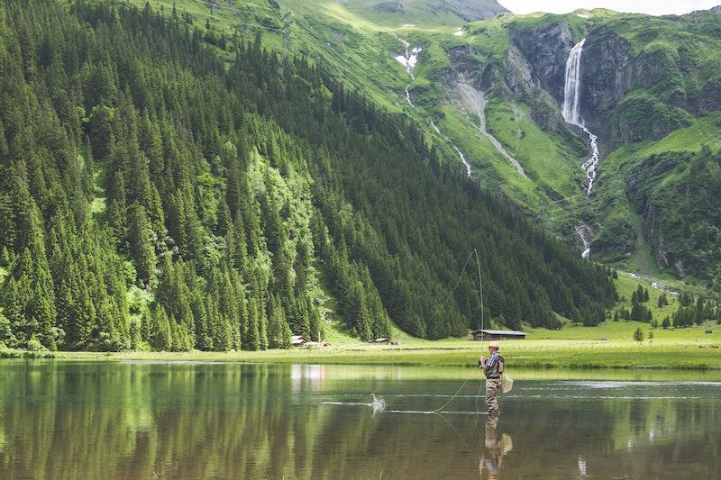 Man Is Fishing in A Lake with A Waterfall in The Background