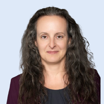 Woman with long, wavy brown hair, wearing a purple blazer and dark blue shirt, smiles.