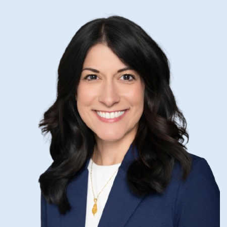 Woman with dark hair smiles, wearing a navy blazer and white shirt, on a light blue background.