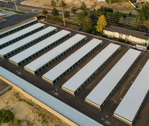 Aerial view of a storage unit facility with rows of individual units, dark asphalt, and a few nearby buildings and a roadway.