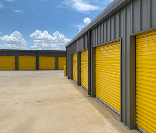 Exterior view of a storage facility with gray metal siding, bright yellow doors, and a blue sky with white clouds.