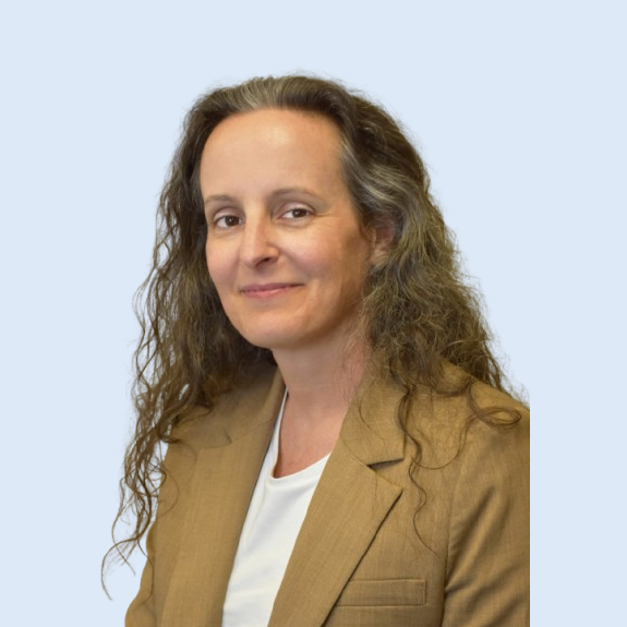 Woman with long, wavy brown hair wearing a tan blazer and white shirt smiles against a light blue background.