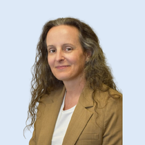 Woman with long, wavy brown hair wearing a tan blazer and white shirt smiles against a light blue background.