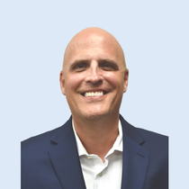 Man in a navy blazer and white shirt smiles. Pale blue background.