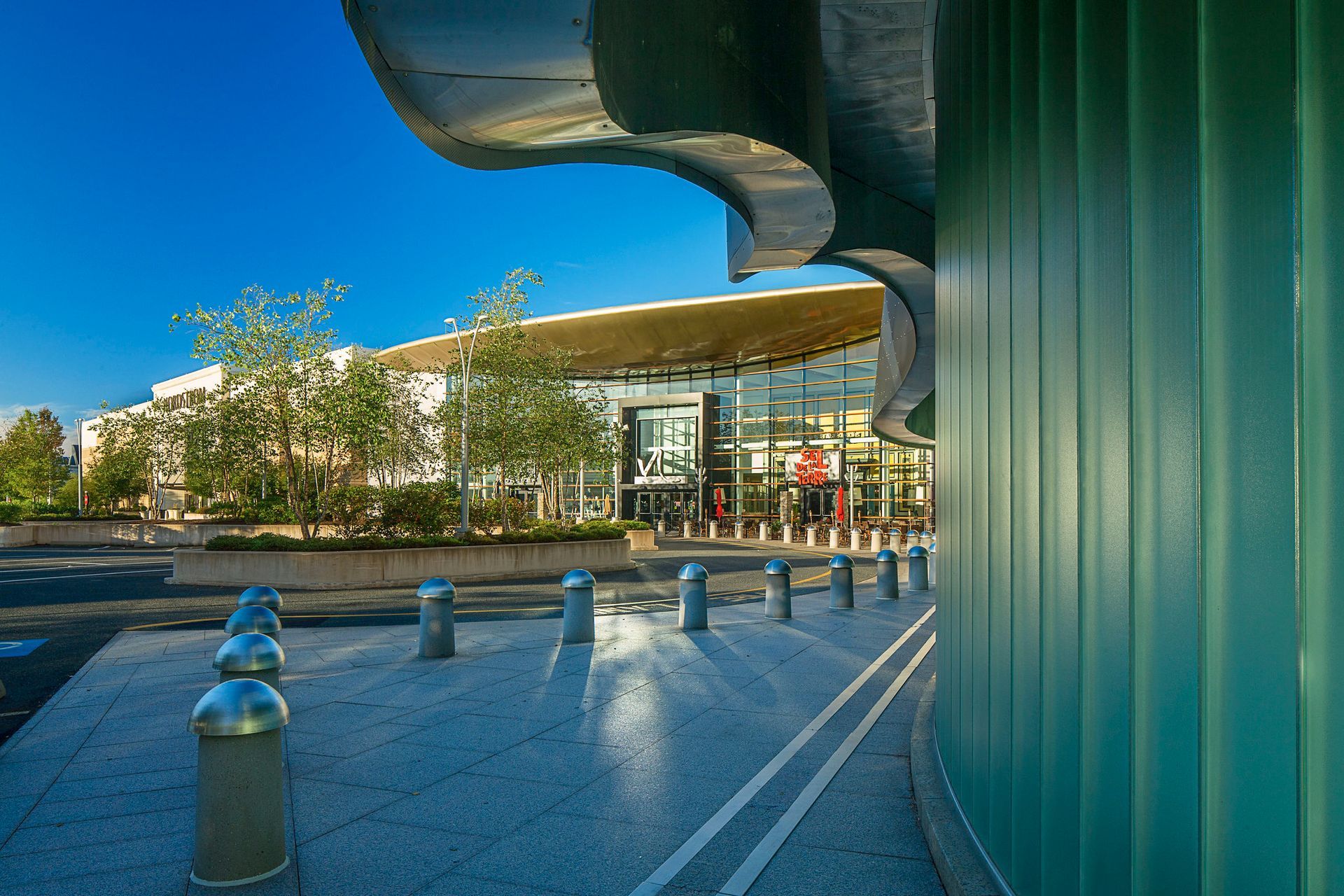 Modern building entrance with green glass wall, curved canopy, and sunlit plaza with trees and bollards