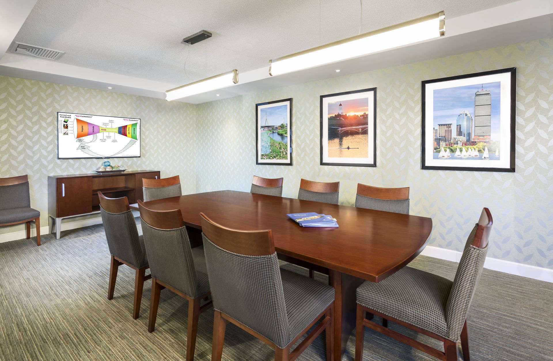 A conference room with a wooden table, chairs, a TV, and three framed pictures on a patterned wall.