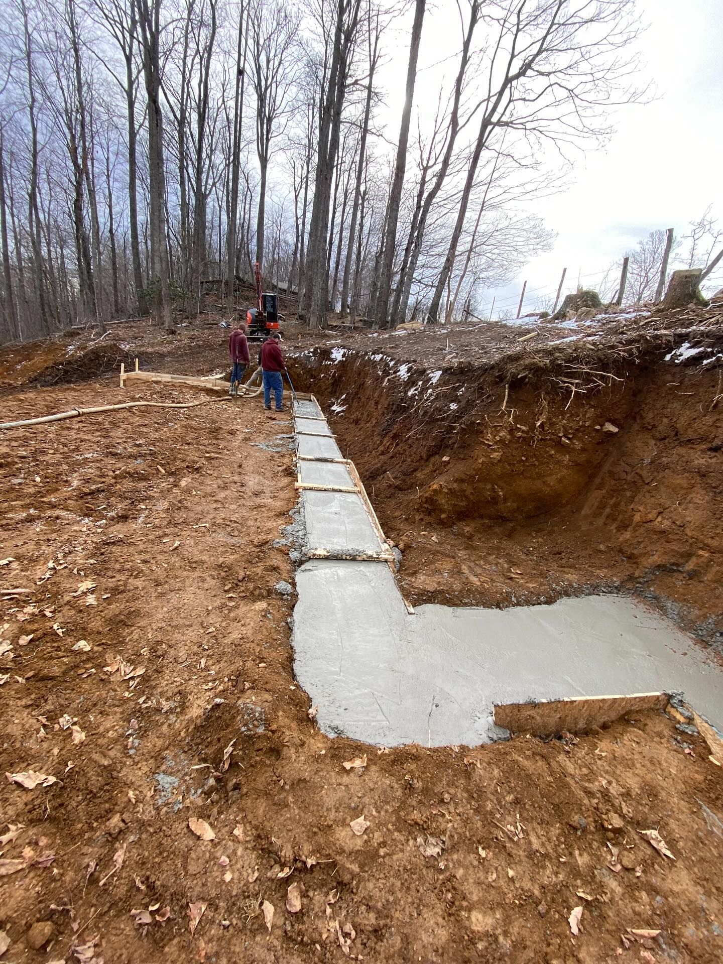 Construction site with concrete foundation poured in an excavated trench, workers present, trees in background.