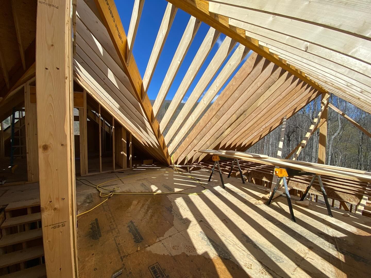 Interior view of a building under construction. Wooden rafters and beams create a roof structure against a bright blue sky.
