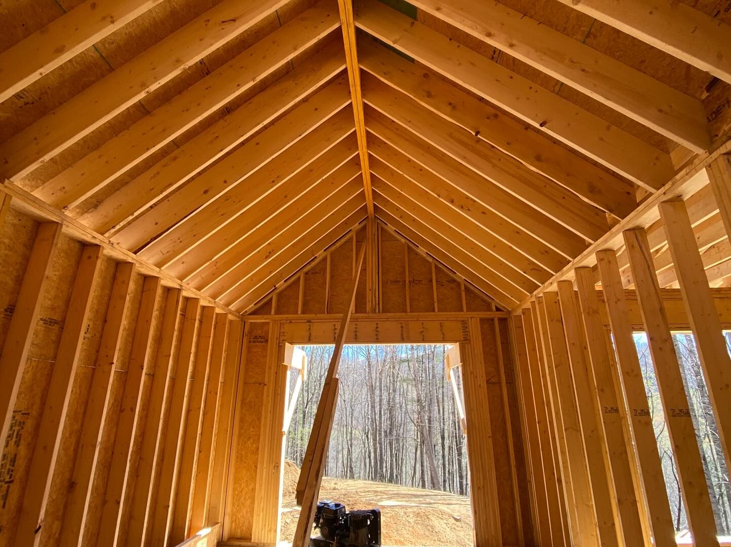 Interior view of a wooden framed building under construction with a gabled roof and doorway opening to trees.