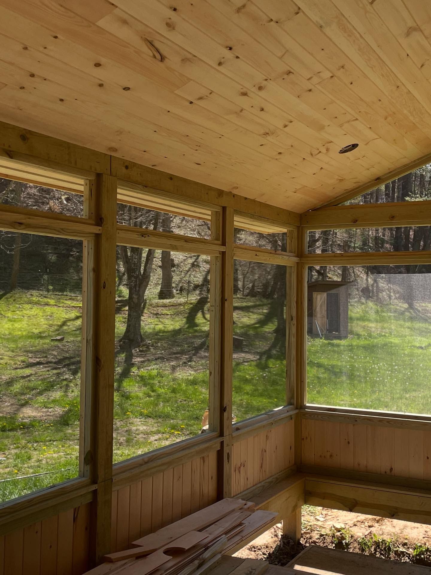 Interior view of a screened porch with wooden framing and paneling, looking out onto a sunny yard.