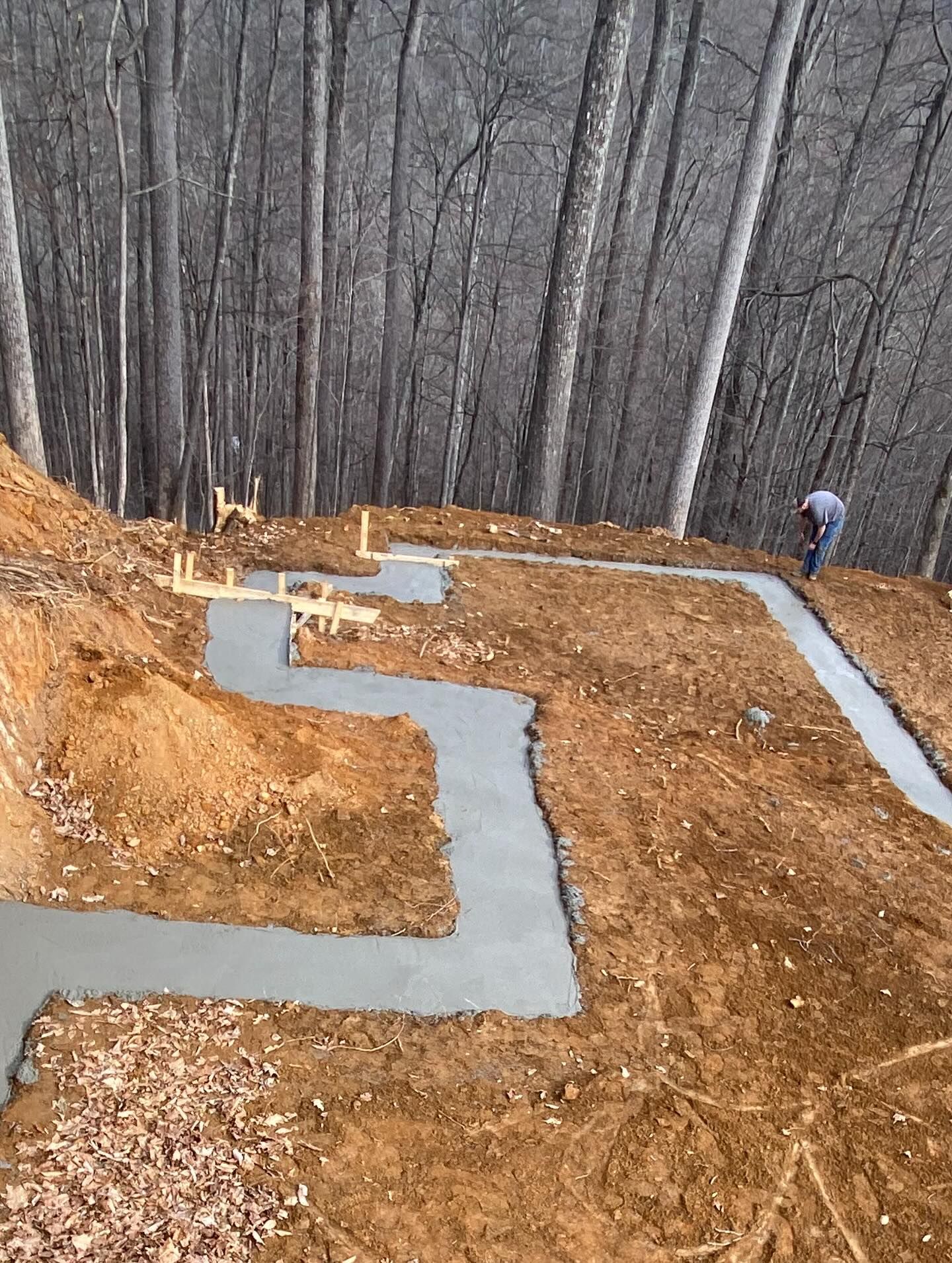 Footings laid in dirt, with worker; woods in background.