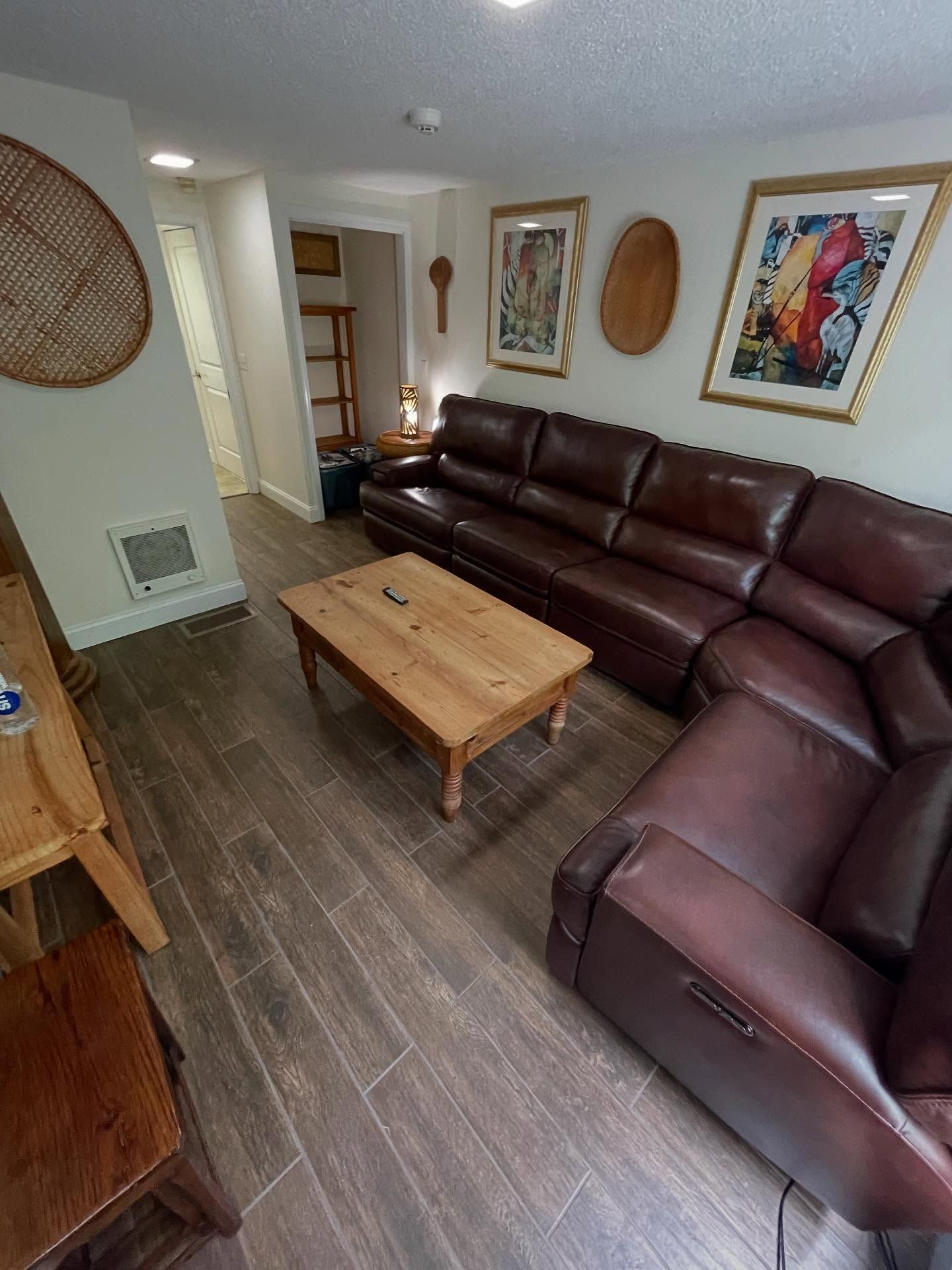Cozy living room with brown leather sectional, wooden coffee table, art, and wood-look flooring.