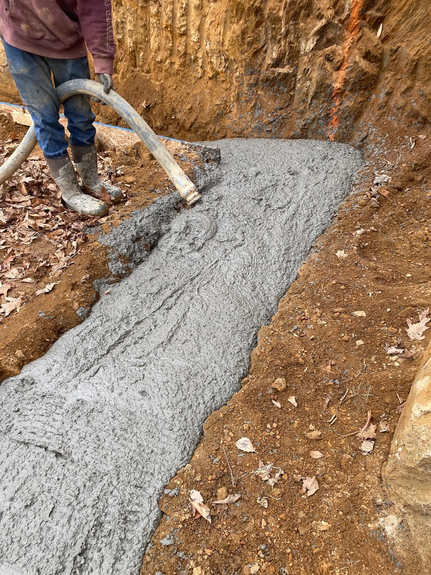 Person pours concrete into a trench. Brown dirt surrounds the gray cement.