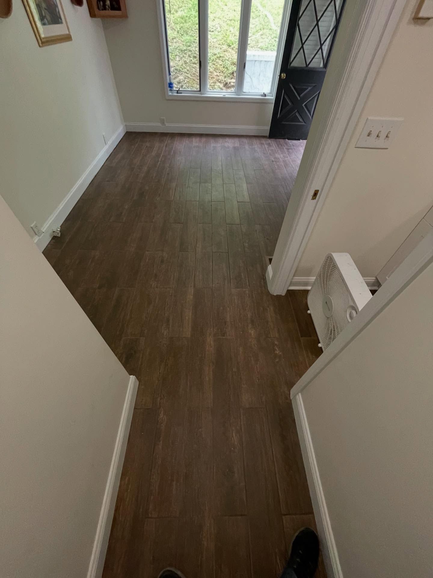 A narrow hallway with wood-look flooring, leading to a room with a window and dark door.