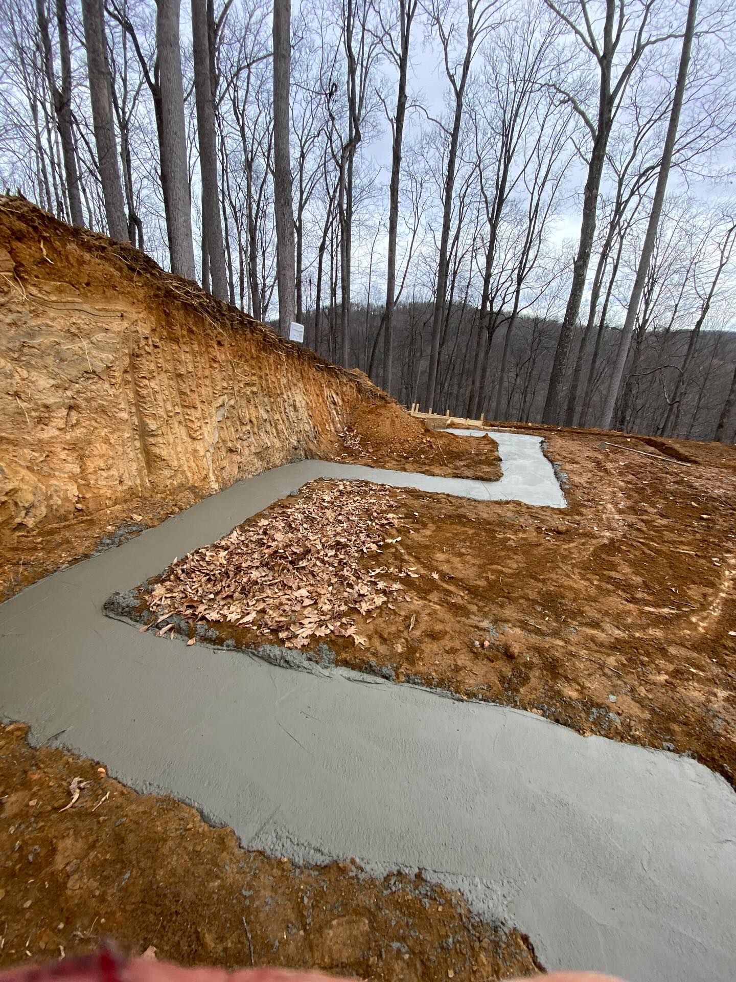 Fresh concrete footings laid on a hillside, forming a winding path. Brown soil and rocks, trees in the background.
