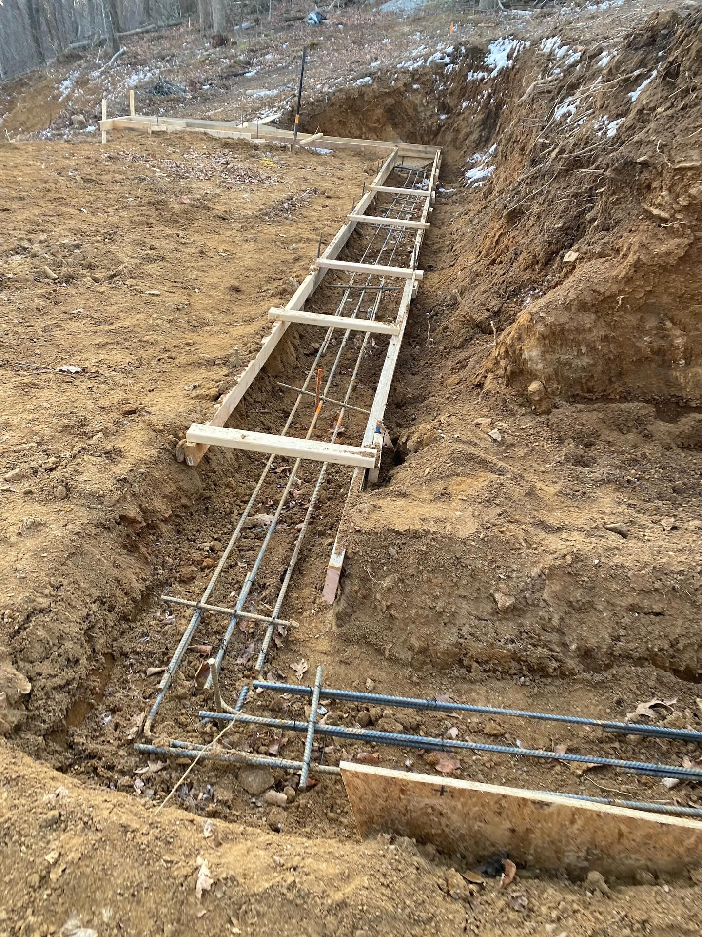 L-shaped foundation with rebar and wooden forms in an excavated trench, ready for concrete, in a dirt setting.