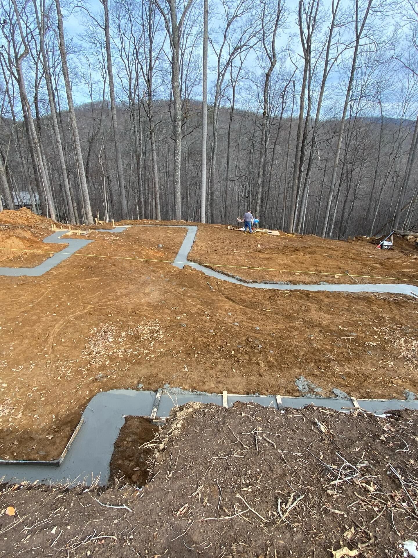 Concrete walkways being constructed on a sloped, brown soil yard with a forest backdrop.