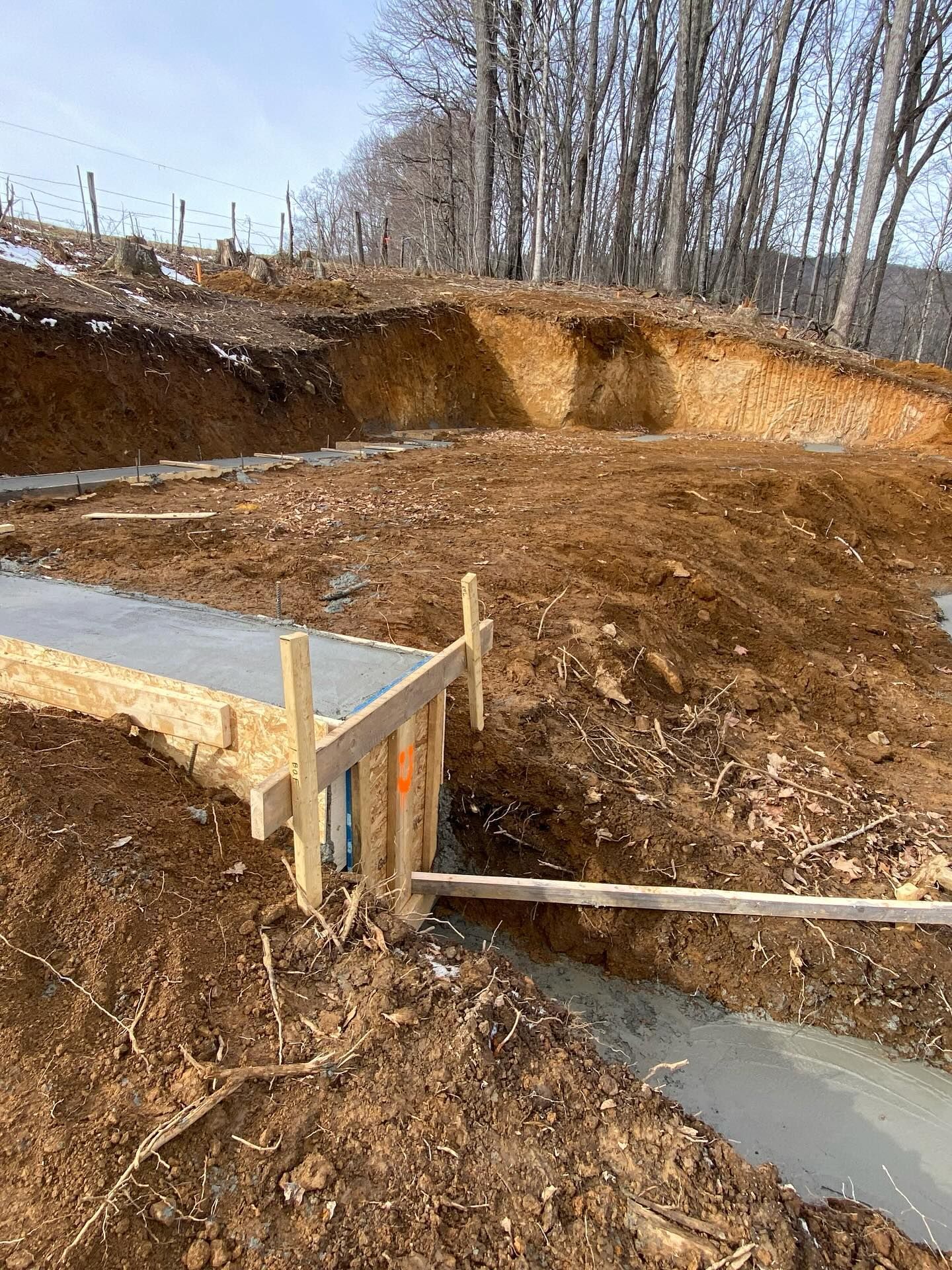 Concrete foundation forms being built on a construction site with excavated dirt and a forested hill in the background.