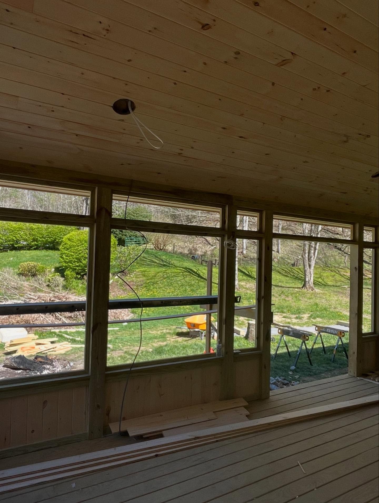 Interior view of a wooden porch with large windows overlooking a green yard and trees. Construction in progress.