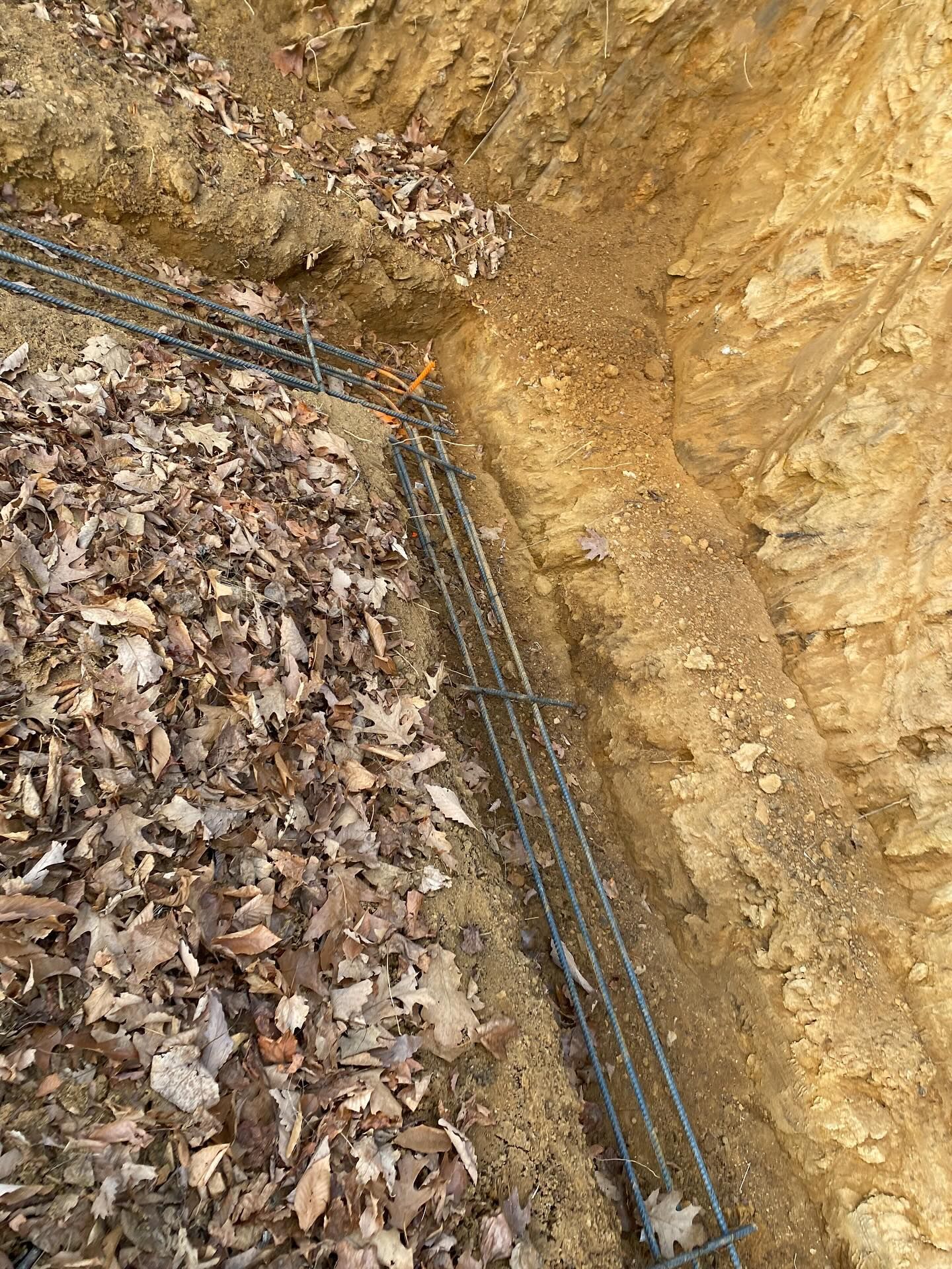 Rebar framework laid in trench, ready for concrete. Brown earth walls with leaves.