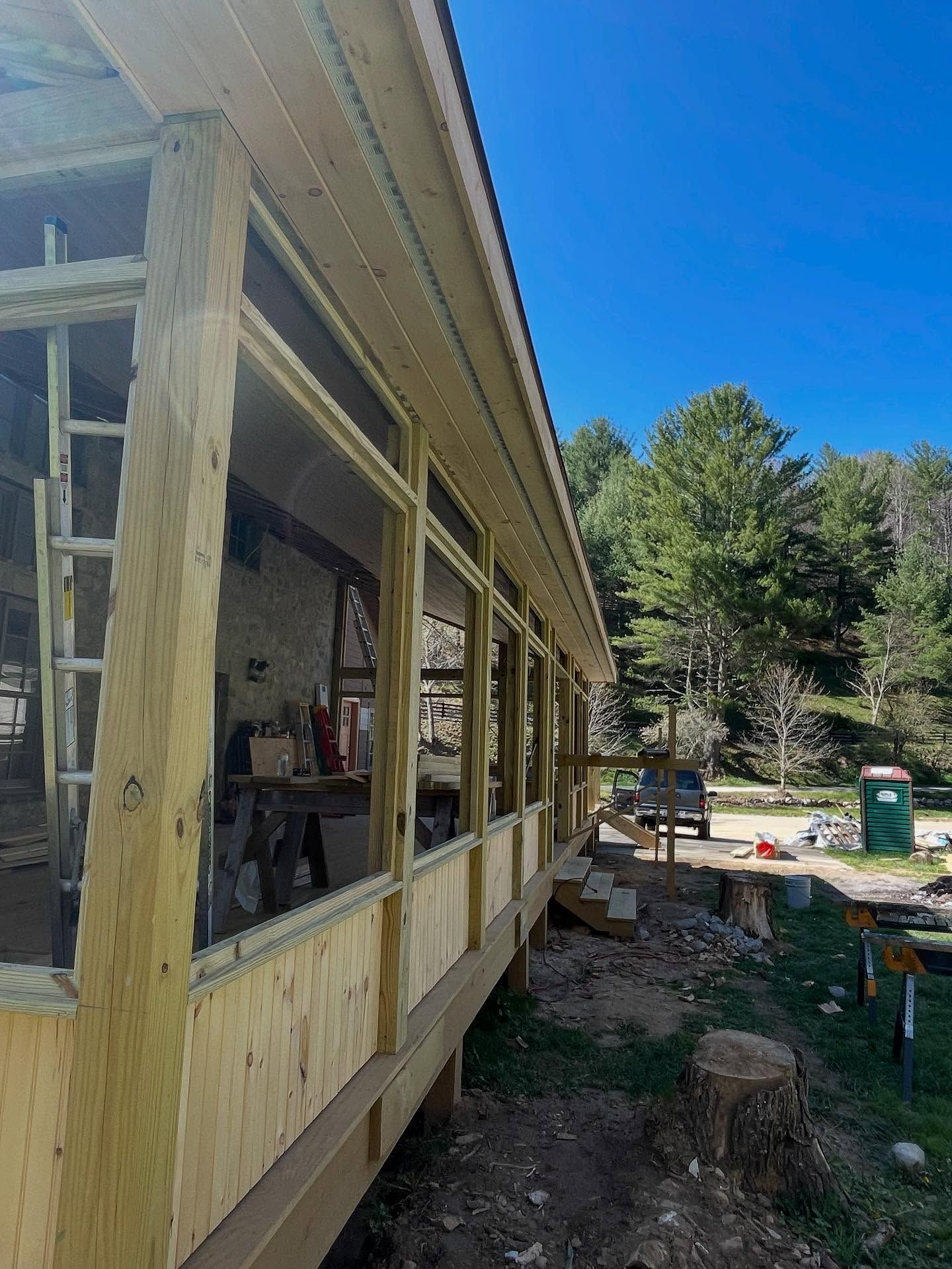 Wooden structure under construction with large windows, a porch, and a tree in the background.