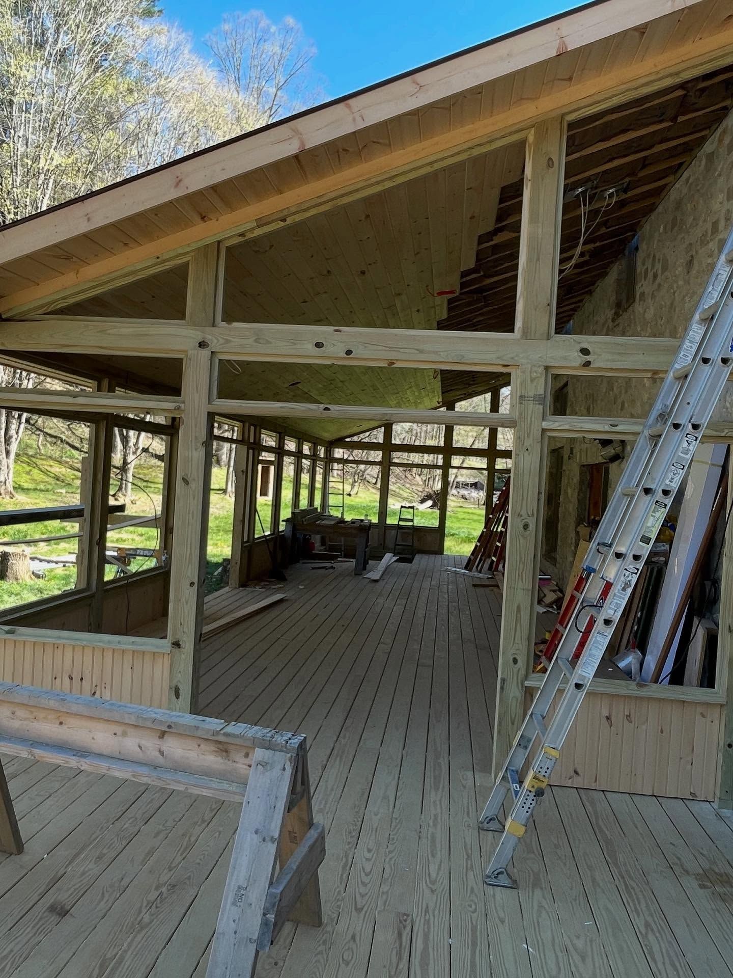 Wooden deck and framework of a screened-in porch under construction, with a ladder and trees visible in the background.