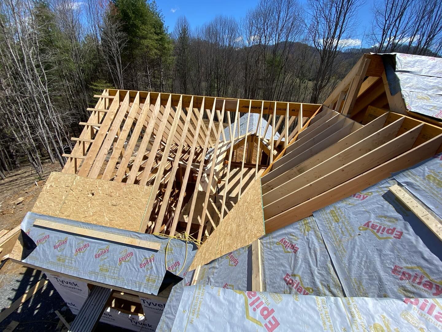 Roof of a house under construction; wooden beams and plywood exposed, covered with silver sheathing, blue sky background.