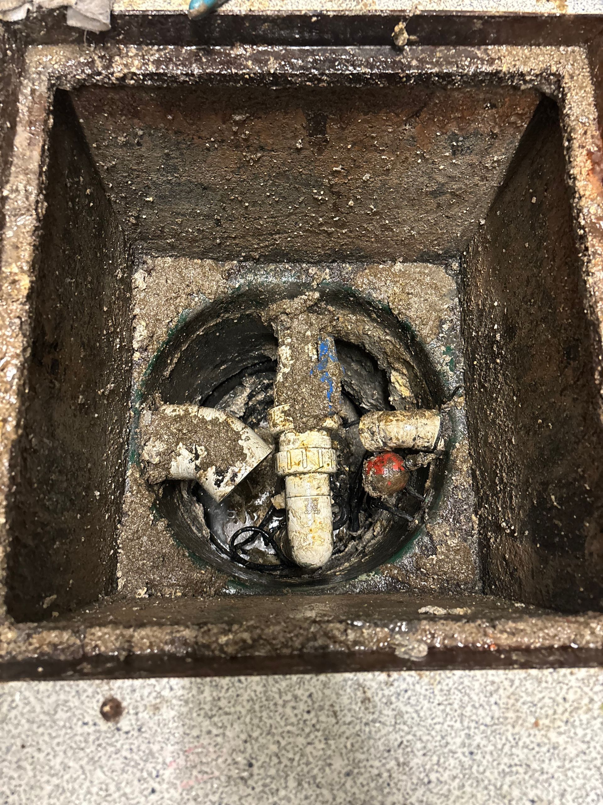A cylindrical, light-colored water heater outside a wooden-sided building, with pipes and a gray foundation.