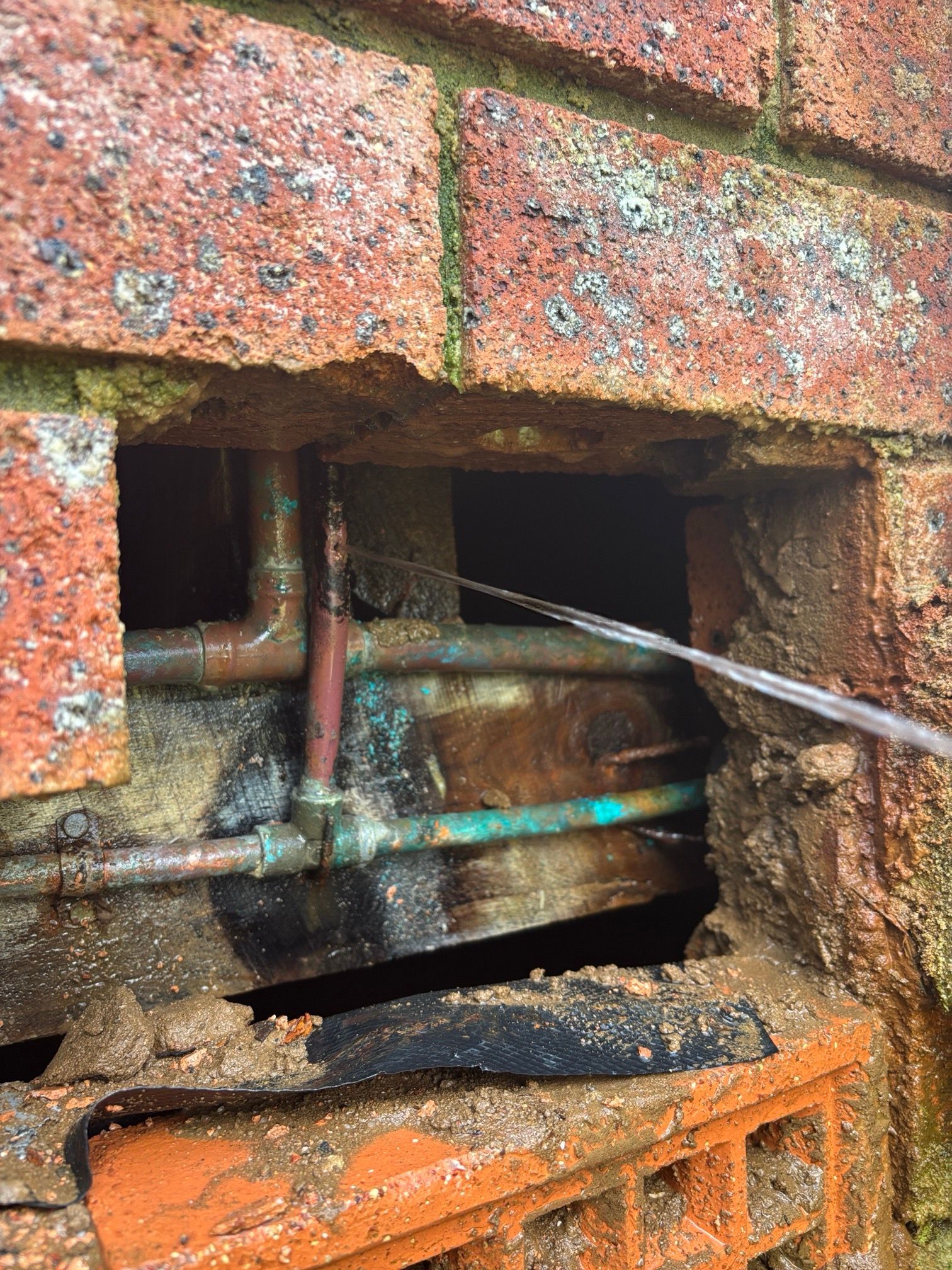 Close-up of brick wall with a hole exposing corroded copper pipes, possibly water or gas lines.