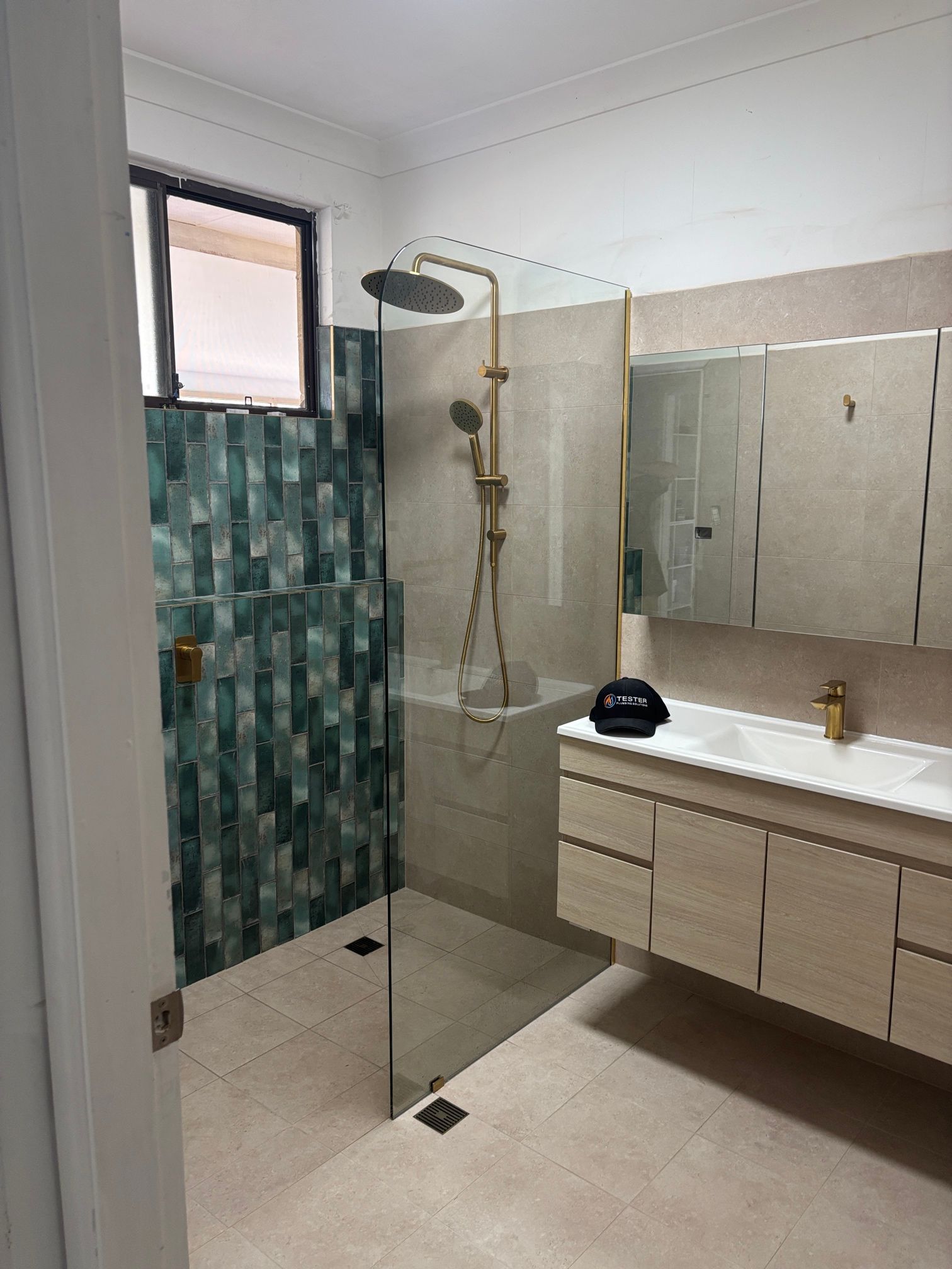 Bathroom with teal tiled shower, gold fixtures, glass shower door, and wood vanity.