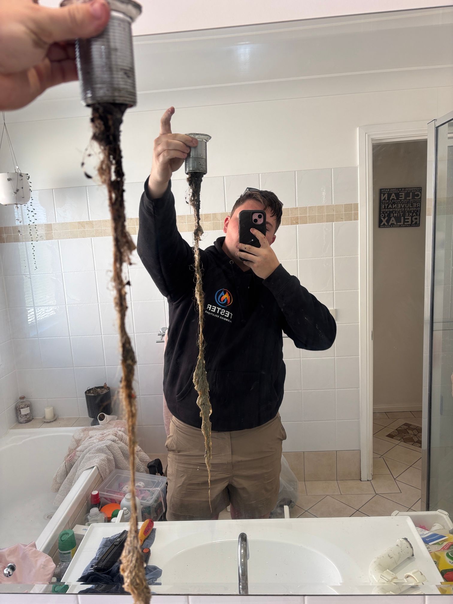 Plumber holding clogged sink drain, standing in a bathroom, reflected in mirror.